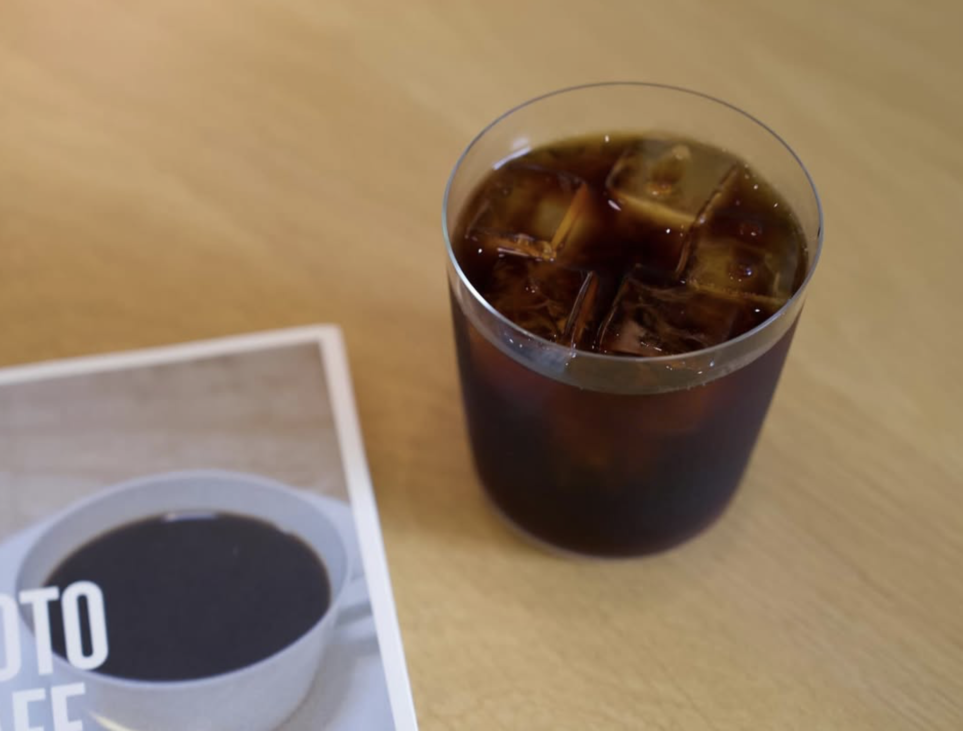 Iced coffee in a clear glass on a light wooden table beside a coffee book